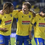 Drie spelers van KVC Westerlo in geel-blauwe wedstrijdkleding vieren samen na een match.