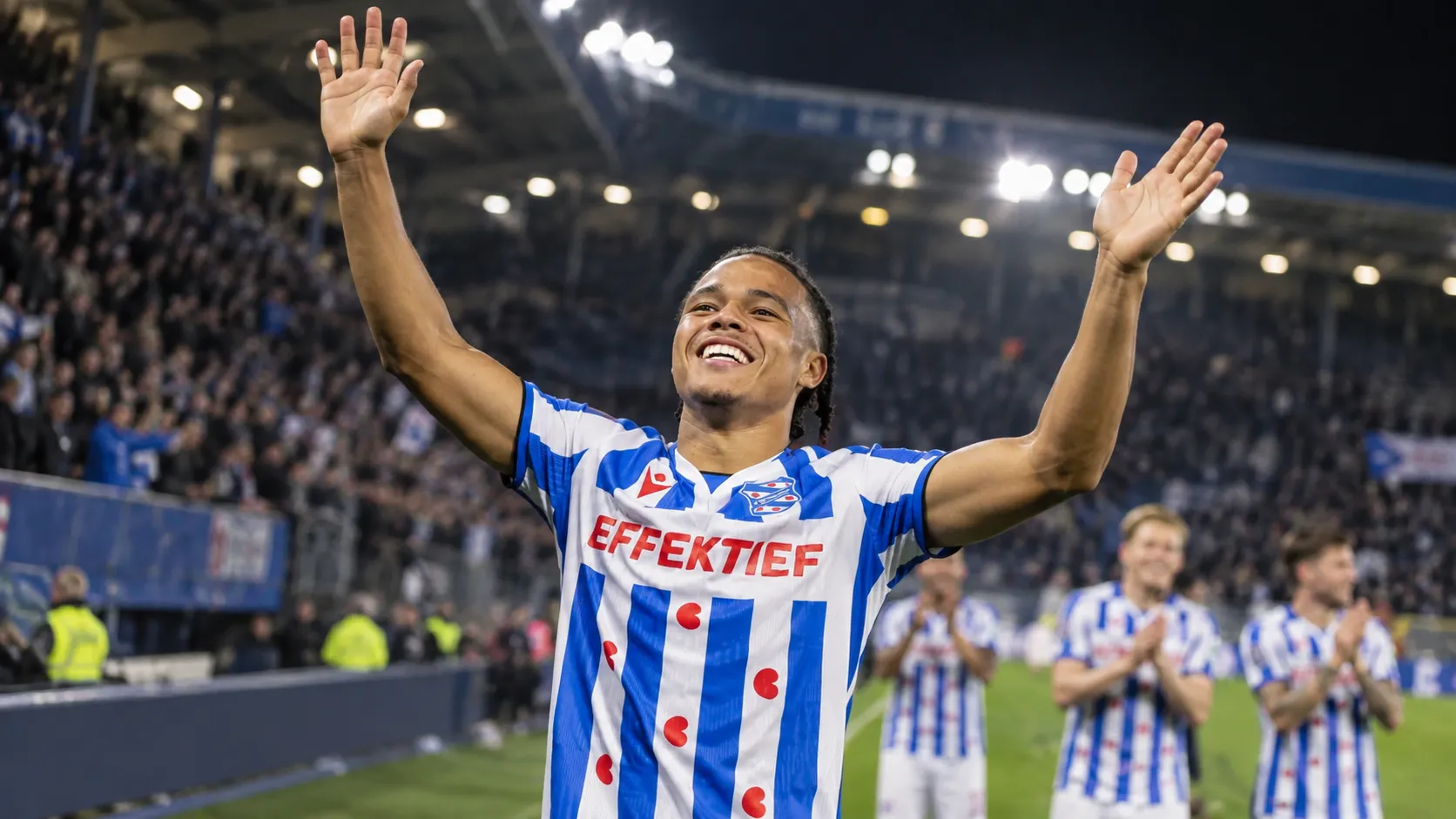 Een speler van sc Heerenveen heft beide armen op in een verlicht stadion.