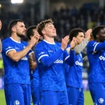 Een groep voetballers in blauwe wedstrijdshirts applaudisseert samen in een stadion.