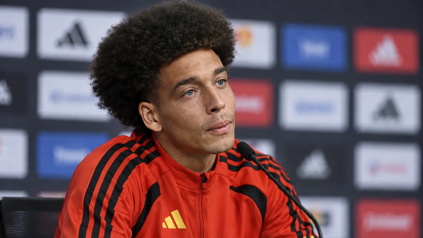 Axel Witsel tijdens een voetbalinterview in een rode sportjas voor een persachtergrond.