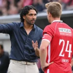 Een coach in donker hemd overlegt langs de zijlijn met een Antwerp-speler in rood tenue.