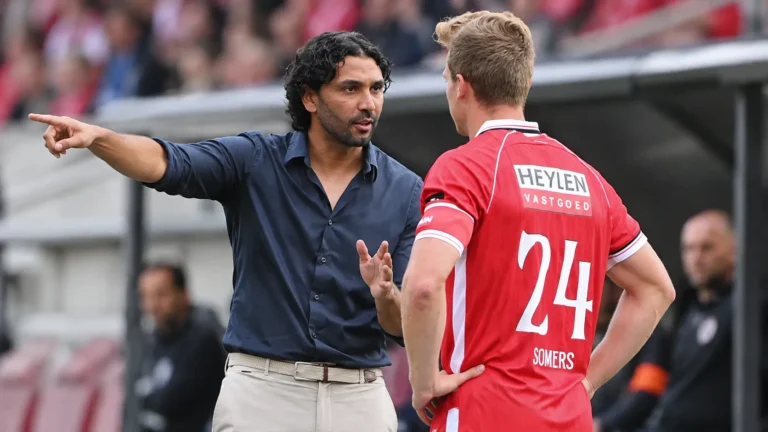 Een coach in donker hemd overlegt langs de zijlijn met een Antwerp-speler in rood tenue.