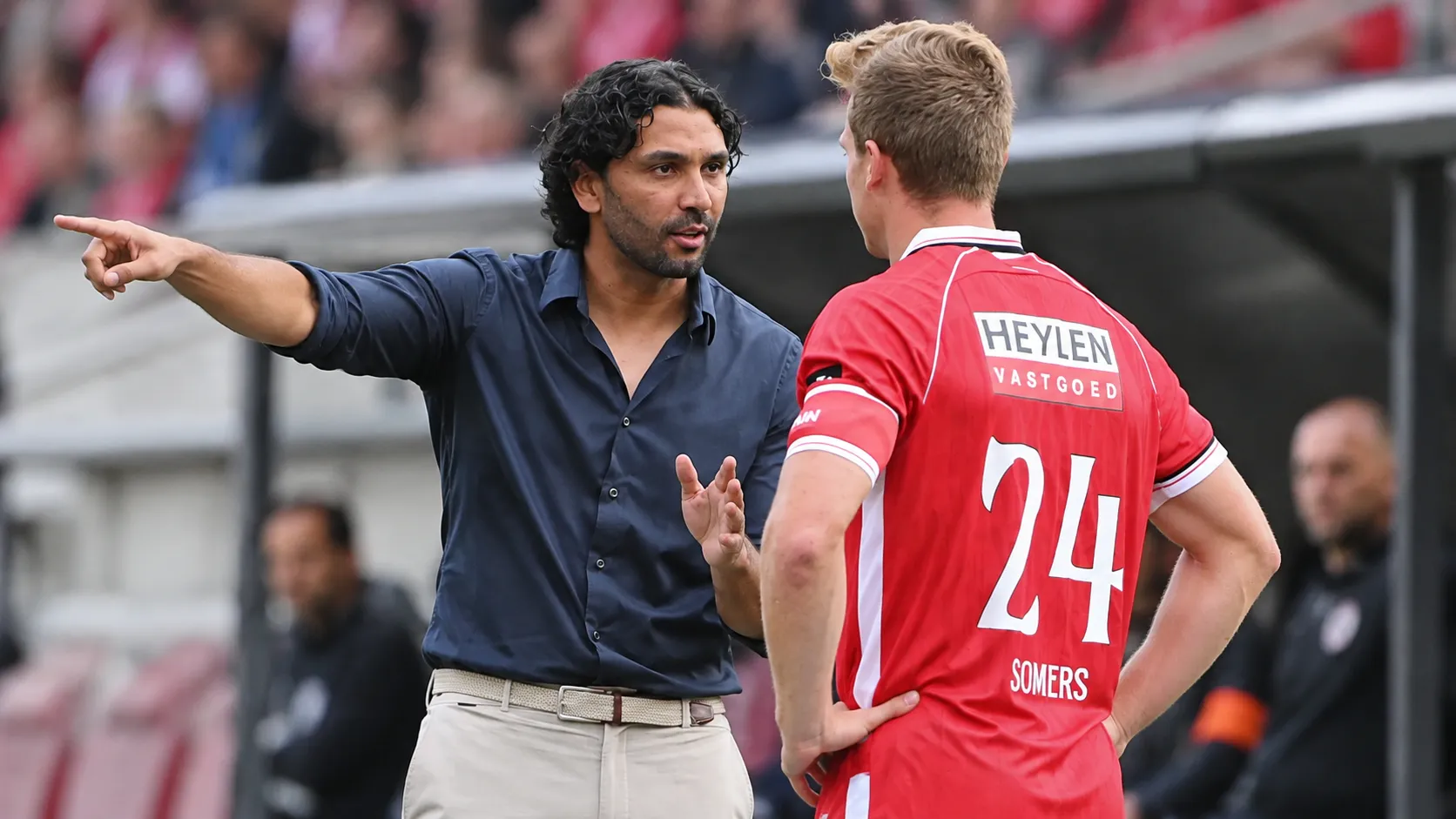 Een coach in donker hemd overlegt langs de zijlijn met een Antwerp-speler in rood tenue.