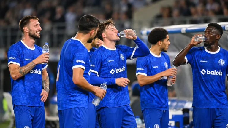 Een groep KAA Gent-spelers in blauwe tenue drinkt water langs het veld.