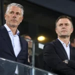 Twee clubbestuurders in donkere kostuums kijken aandachtig toe vanaf de stadiontribune.