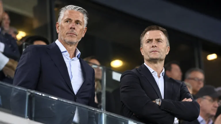 Twee clubbestuurders in donkere kostuums kijken aandachtig toe vanaf de stadiontribune.