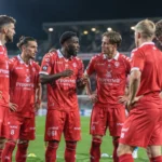 Een groep voetballers in rode wedstrijdtenues vormt een kring op een verlicht veld.