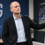 Een coach van KRC Genk staat in een tactische vergaderruimte bij een vast clubembleem aan de muur.