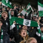 Supporters van La Louvière zwaaien met groen-witte vlaggen in de tribune.