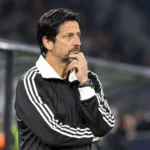 Een coach in een zwart-wit trainingsjack volgt geconcentreerd de wedstrijd vanaf de zijlijn.