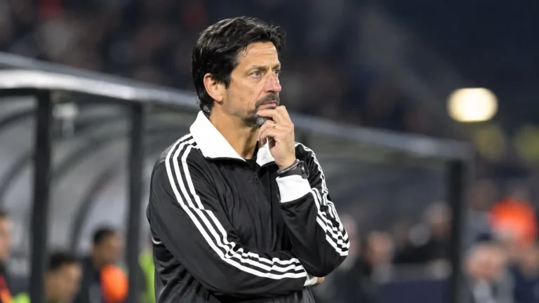 Een coach in een zwart-wit trainingsjack volgt geconcentreerd de wedstrijd vanaf de zijlijn.