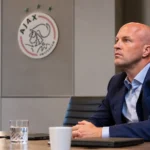 Een clubofficial in blauw pak zit in een Ajax-vergaderruimte met een Ajax-embleem op de wand.