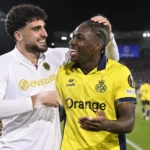 Twee voetballers in gele en witte shirts vieren een moment op het veld onder stadionverlichting.