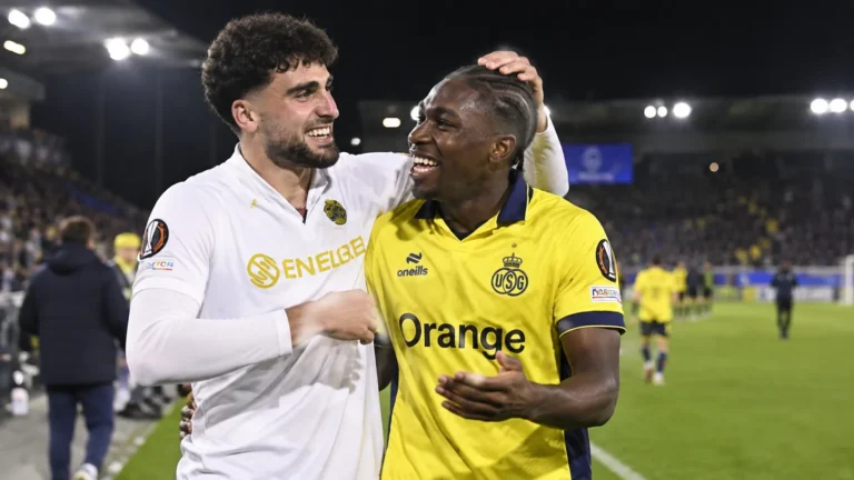 Twee voetballers in gele en witte shirts vieren een moment op het veld onder stadionverlichting.