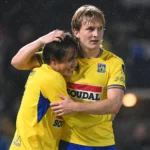 Twee Westerlo-spelers in geel-blauwe wedstrijdkleding vieren samen een moment in de regen.