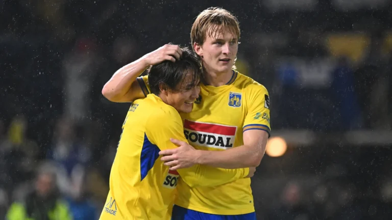Twee Westerlo-spelers in geel-blauwe wedstrijdkleding vieren samen een moment in de regen.