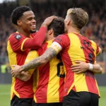 Drie voetballers in rood-geel tenue vieren samen een moment op het veld.