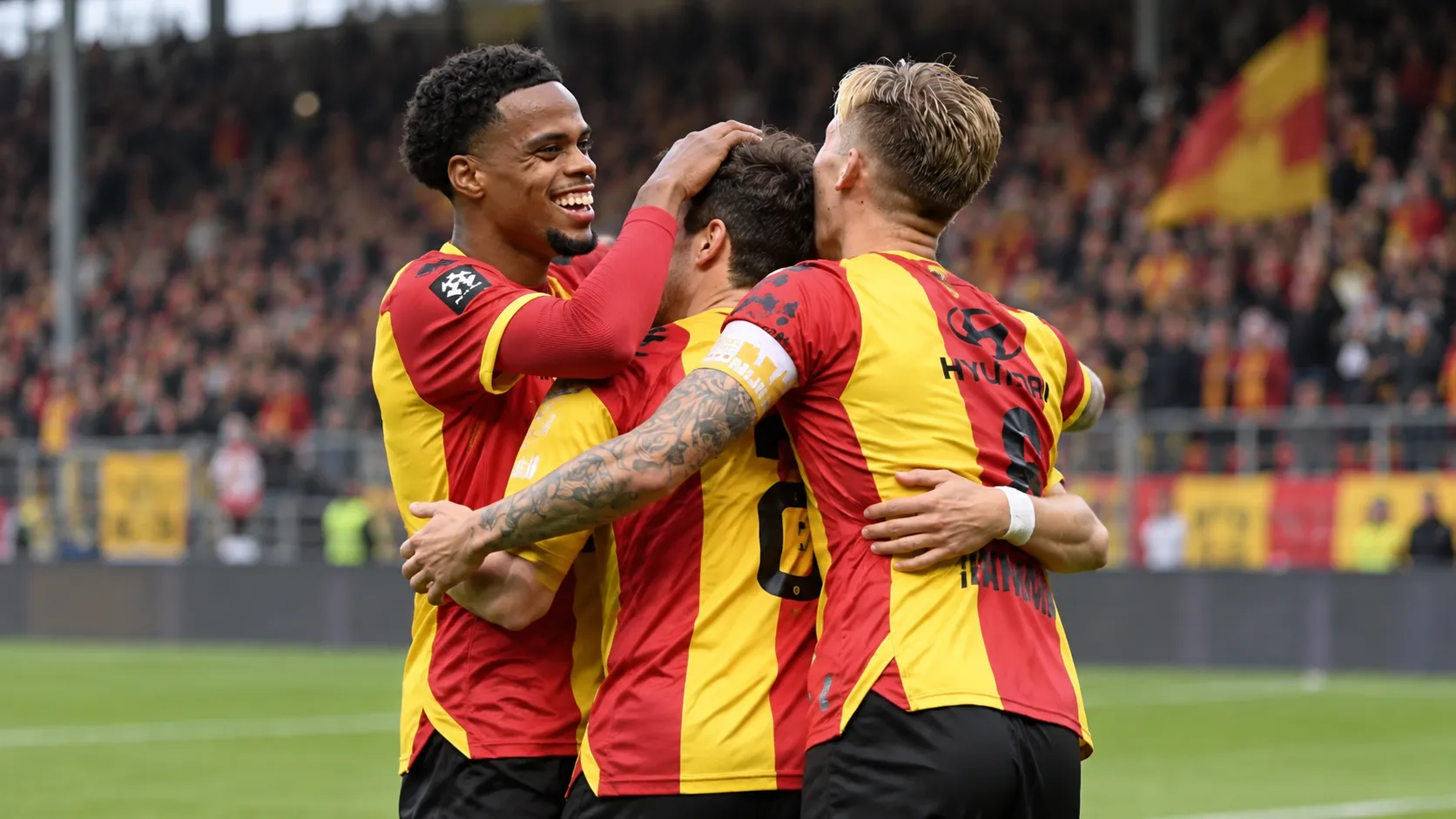 Drie voetballers in rood-geel tenue vieren samen een moment op het veld.