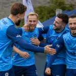 Een groep voetballers in blauwe shirts viert samen langs het veld.