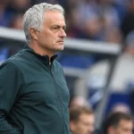 Een grijs­harige coach in een donker trainingsjack langs de zijlijn van een voetbalstadion.
