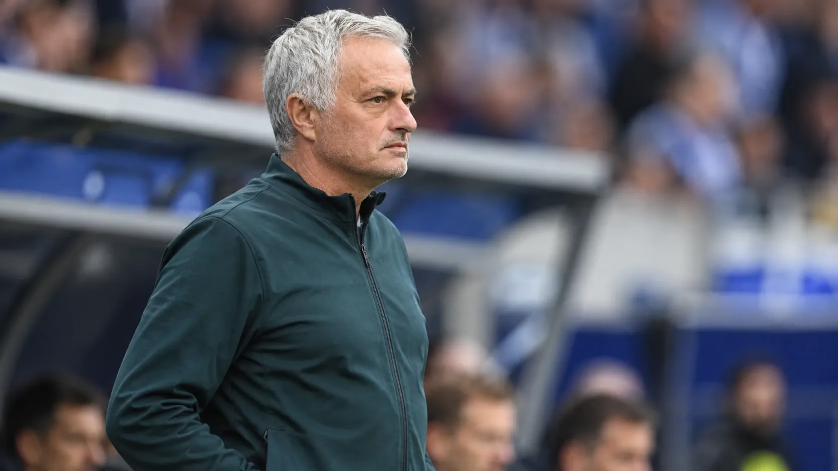 Een grijsharige coach in een donker trainingsjack langs de zijlijn van een voetbalstadion.