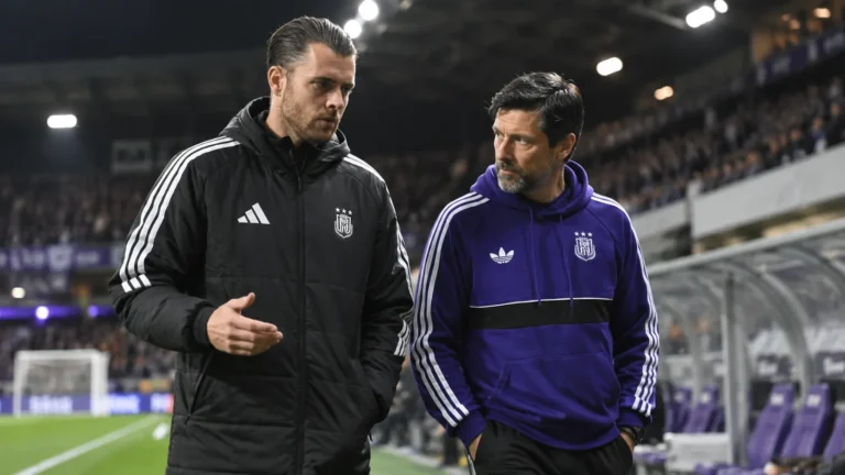 Twee stafleden van Anderlecht wandelen langs de zijlijn van een verlicht stadion.