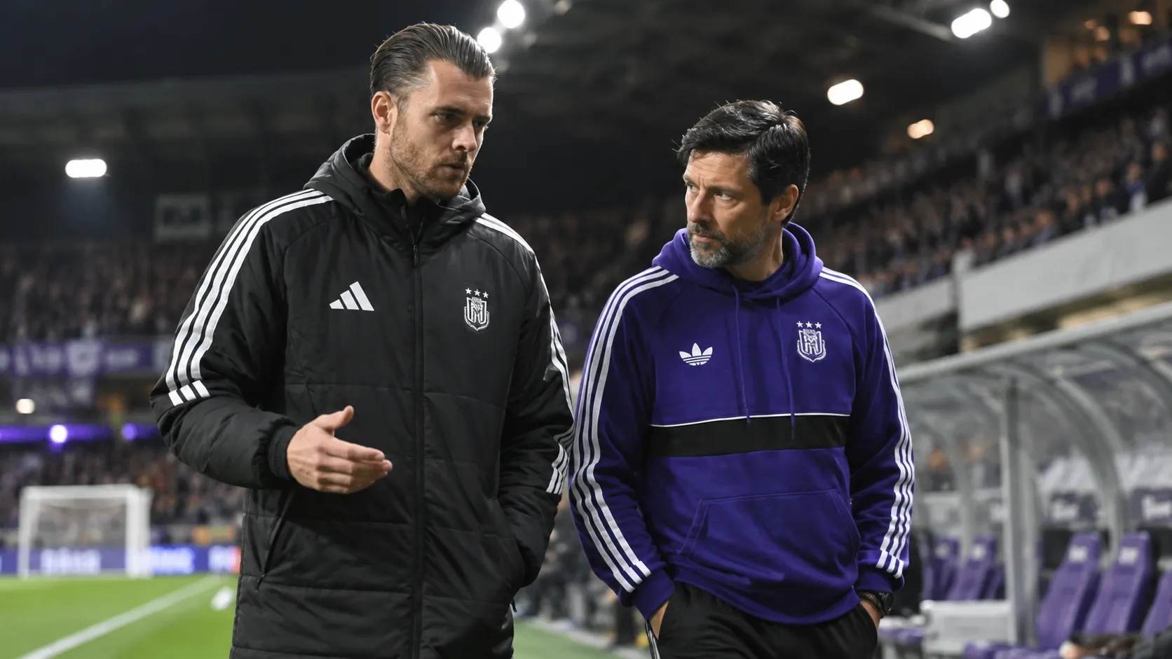 Twee stafleden van Anderlecht wandelen langs de zijlijn van een verlicht stadion.