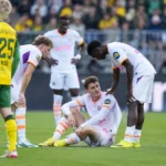 Spelers staan rond een zittende ploegmaat tijdens een spelonderbreking op het veld.