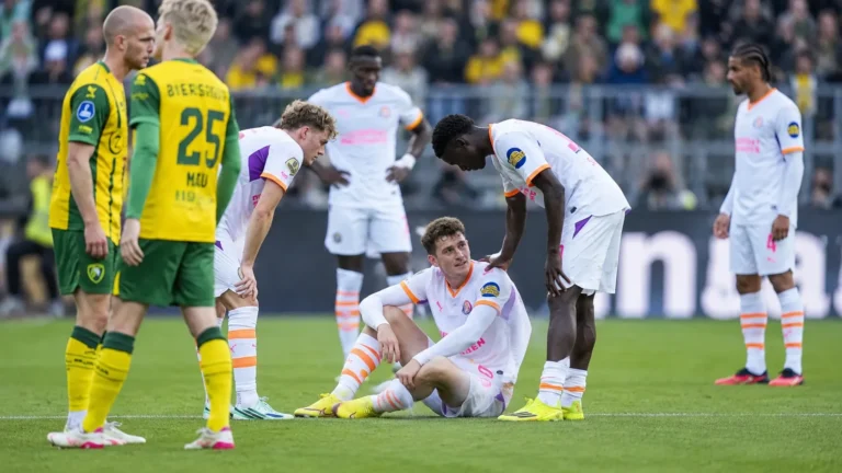 Spelers staan rond een zittende ploegmaat tijdens een spelonderbreking op het veld.