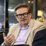 Een clubofficial in een beige colbert spreekt in de VIP-zone van een voetbalstadion.