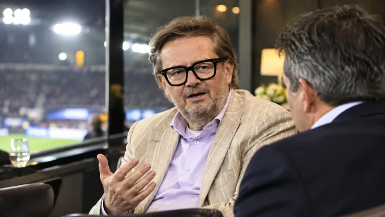 Een clubofficial in een beige colbert spreekt in de VIP-zone van een voetbalstadion.