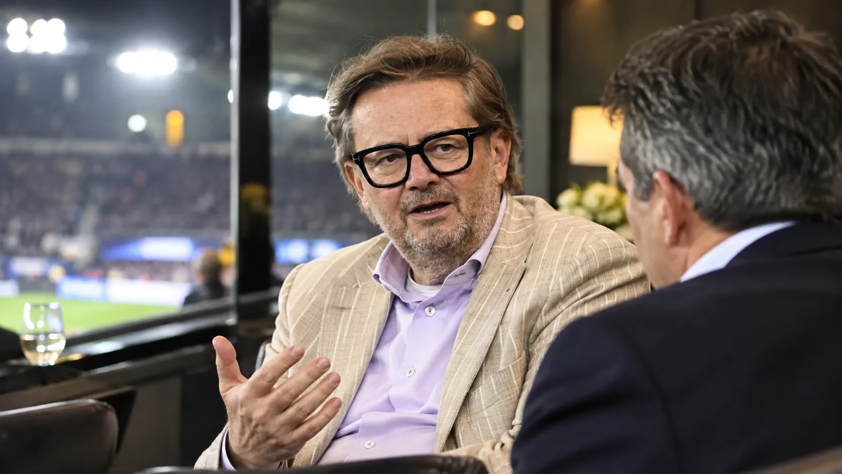 Een clubofficial in een beige colbert spreekt in de VIP-zone van een voetbalstadion.
