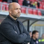 Een niet-spelende stafmedewerker in een zwarte jas naast de technische zone van de Freethiel.