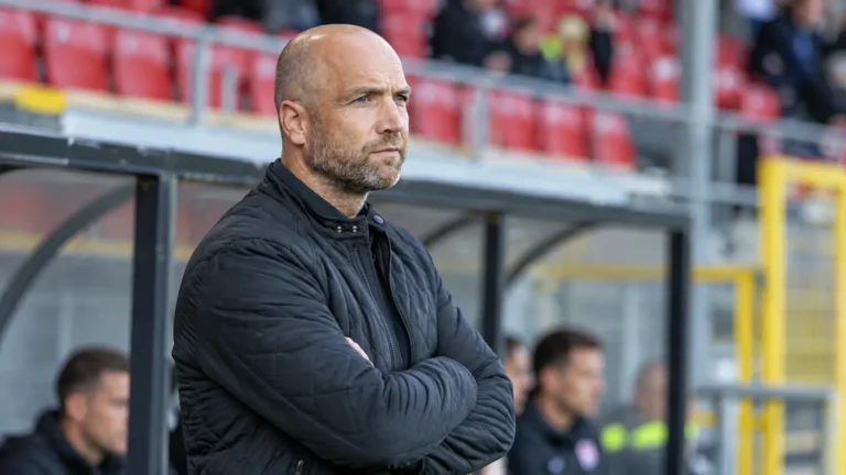 Een niet-spelende stafmedewerker in een zwarte jas naast de technische zone van de Freethiel.