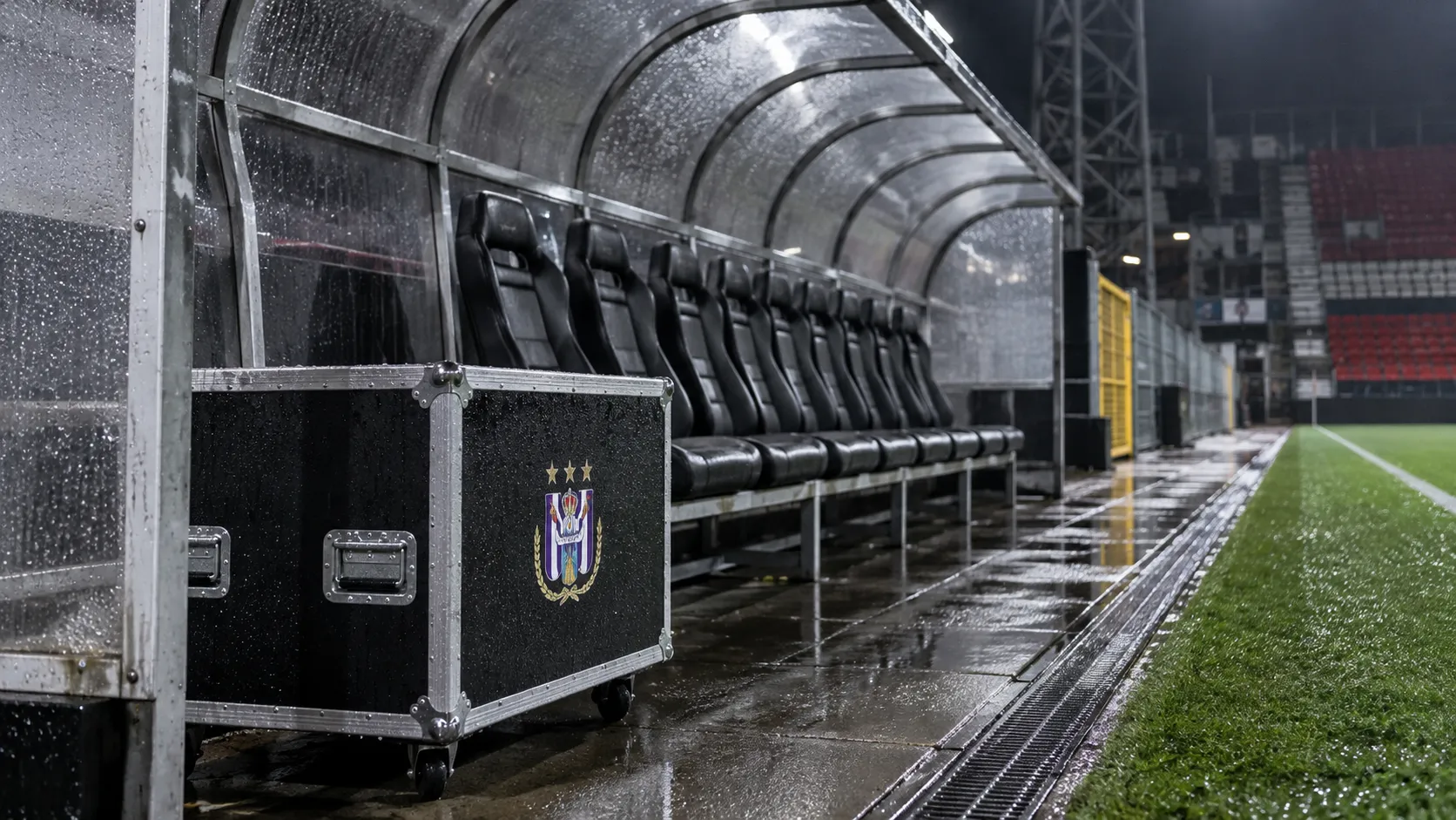 Een lege bezoekersdug-out met een materiaaltrunk van Anderlecht naast het veld.