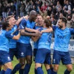 Een groep voetballers in lichtblauwe tenues viert voor een juichende thuistribune.