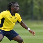 Massiré Sylla in geel en donkerblauw tenue tijdens een verdedigend spelfragment op een grasveld.