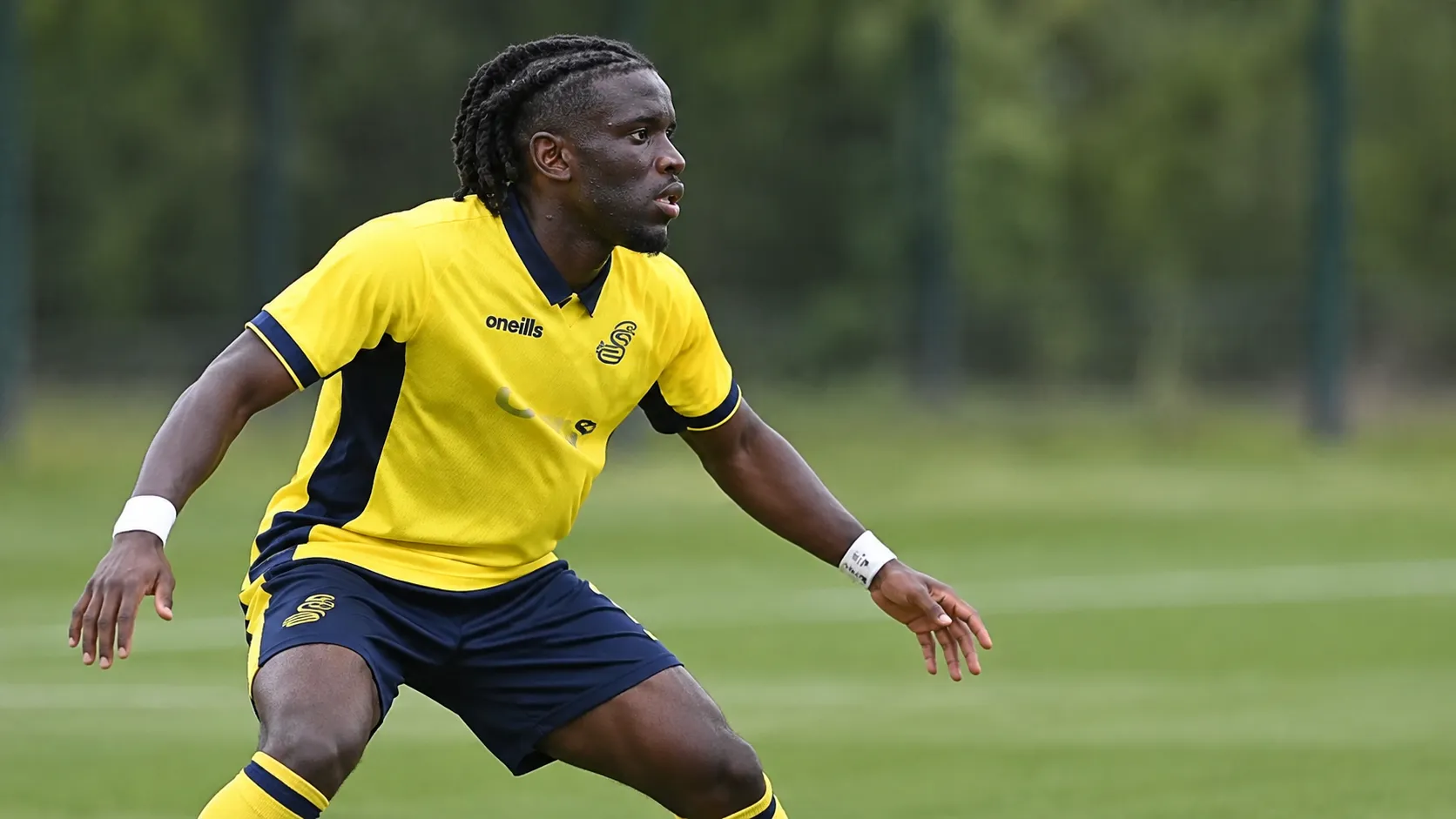 Massiré Sylla in geel en donkerblauw tenue tijdens een verdedigend spelfragment op een grasveld.