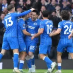Een groep voetballers in blauw gestreepte shirts viert samen op het veld voor een volle tribune.