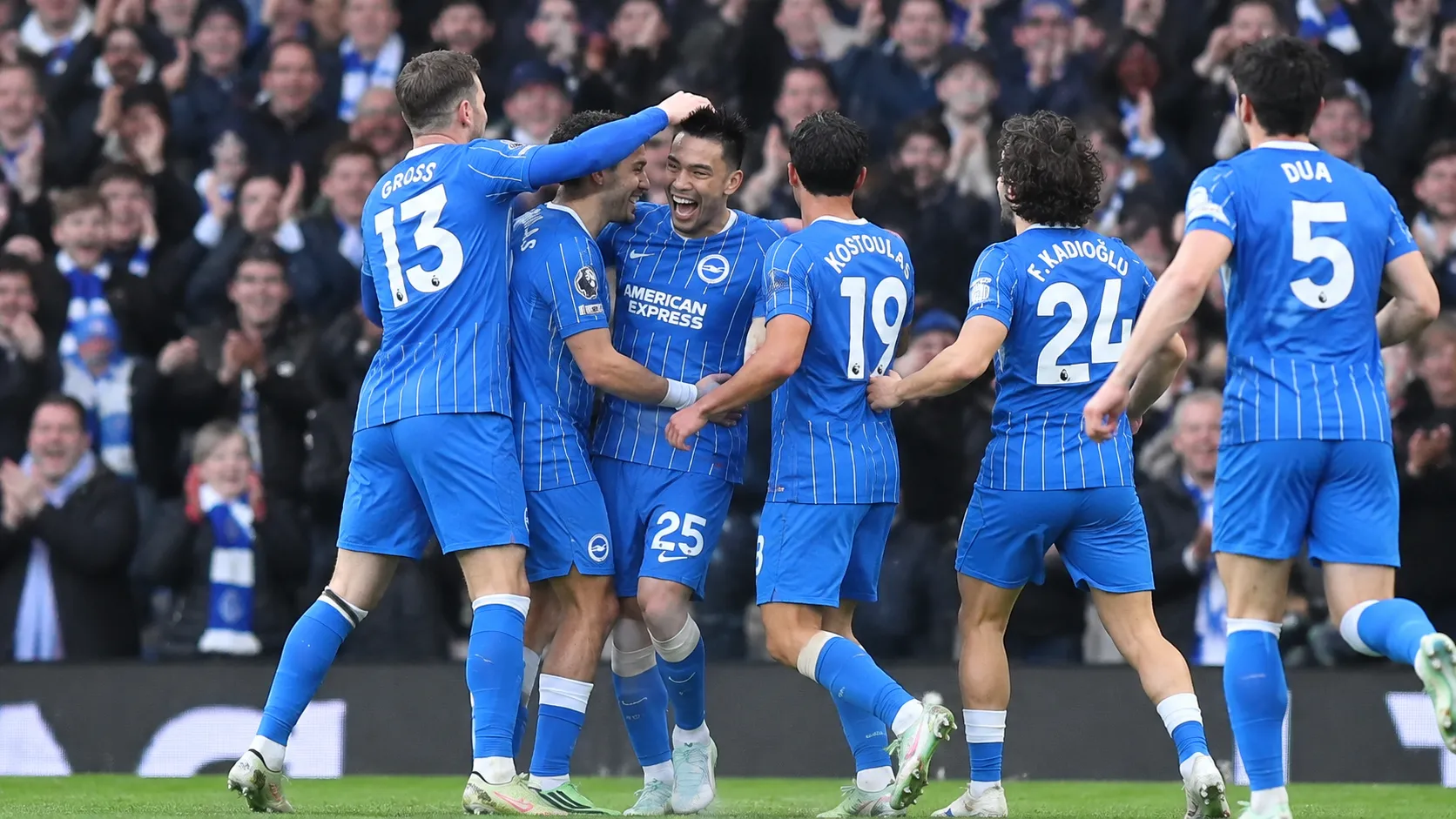 Een groep voetballers in blauw gestreepte shirts viert samen op het veld voor een volle tribune.