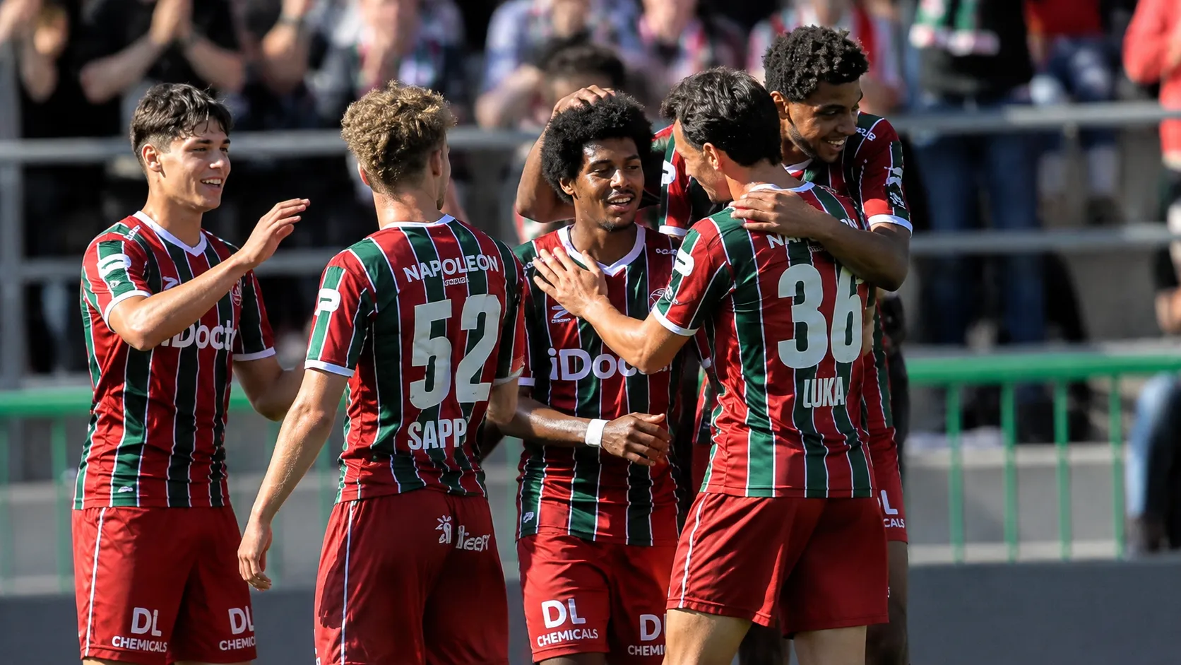 Een groep voetballers in rood-groen gestreepte tenues viert samen naast het veld.