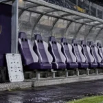 Lege bezoekersdug-out met een klein RSC Anderlecht-embleem op een vast wandpaneel.