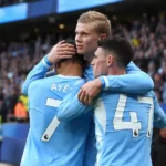 Drie voetballers in lichtblauwe shirts omhelzen elkaar naast het veld.