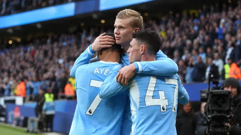 Drie voetballers in lichtblauwe shirts omhelzen elkaar naast het veld.