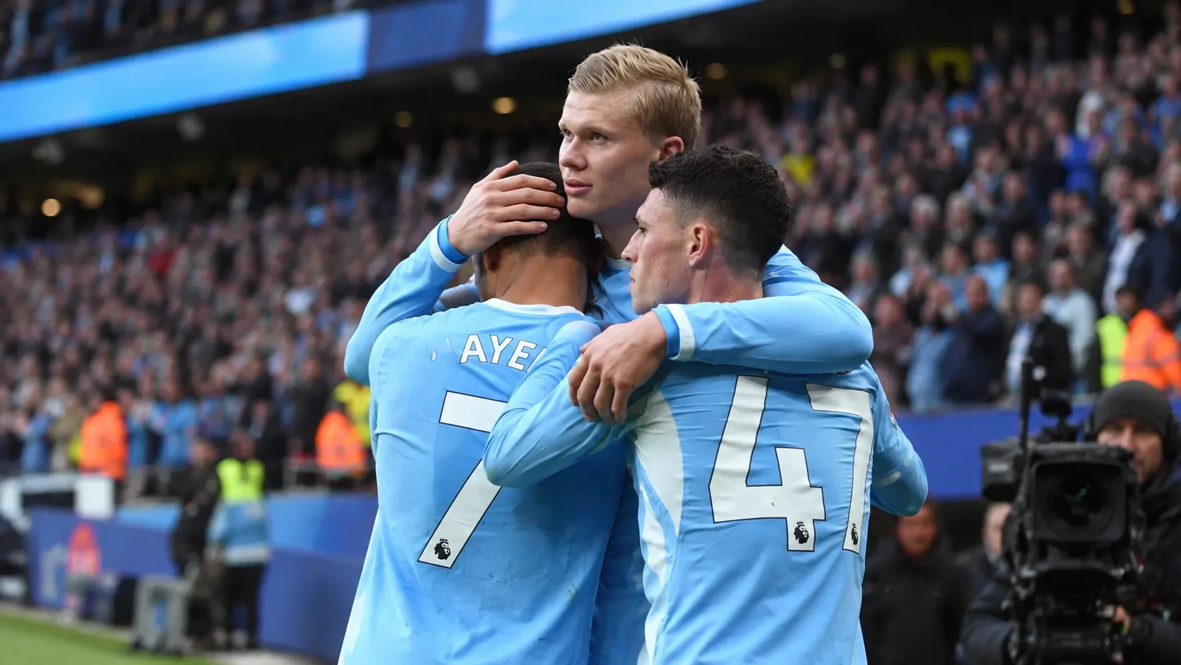 Drie voetballers in lichtblauwe shirts omhelzen elkaar naast het veld.