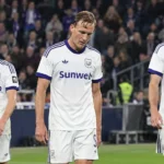 Drie voetballers in witte shirts verlaten zichtbaar ontgoocheld het veld na een wedstrijd.