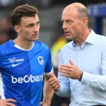 Een voetballer in een blauw shirt overlegt langs de zijlijn met een staflid.