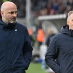 Twee niet-spelende clubvertegenwoordigers in donkere jassen naast de zijlijn van een stadion.