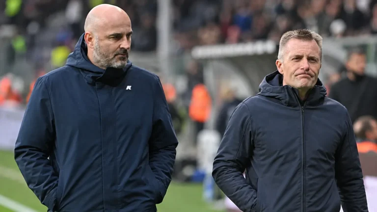 Twee niet-spelende clubvertegenwoordigers in donkere jassen naast de zijlijn van een stadion.