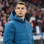 Cristiano Ronaldo in een blauwe jas langs de zijlijn van een voetbalstadion.